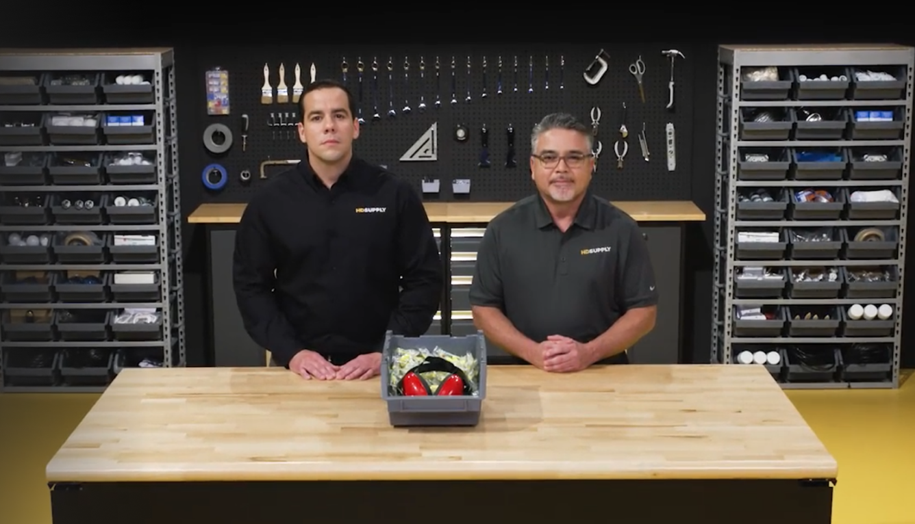 Two HD Supply trainers standing side-by-side inside of a maintenance shop and behind a wooden table with a safety headphones inside a black box placed in the center